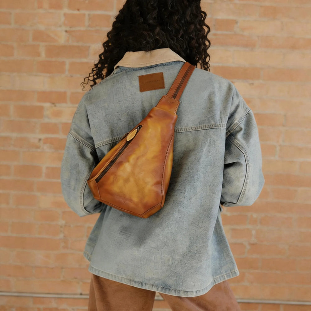 Person wearing a denim jacket with a brown leather bag over one shoulder against a brick wall.
