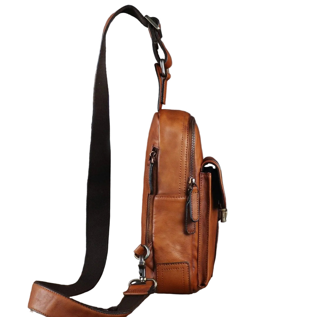 A brown leather crossbody bag with multiple zippered pockets and a long adjustable strap, displayed upright on a vintage-style wooden trunk against a dark gray textured backdrop.