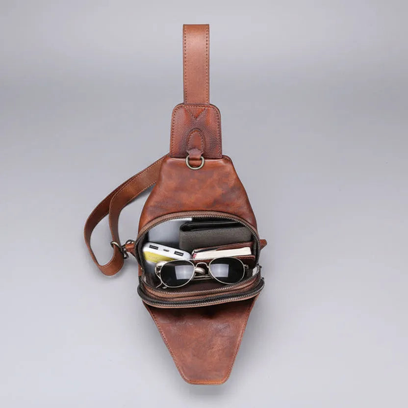 Brown leather sling bag with sunglasses and phone on a gray background