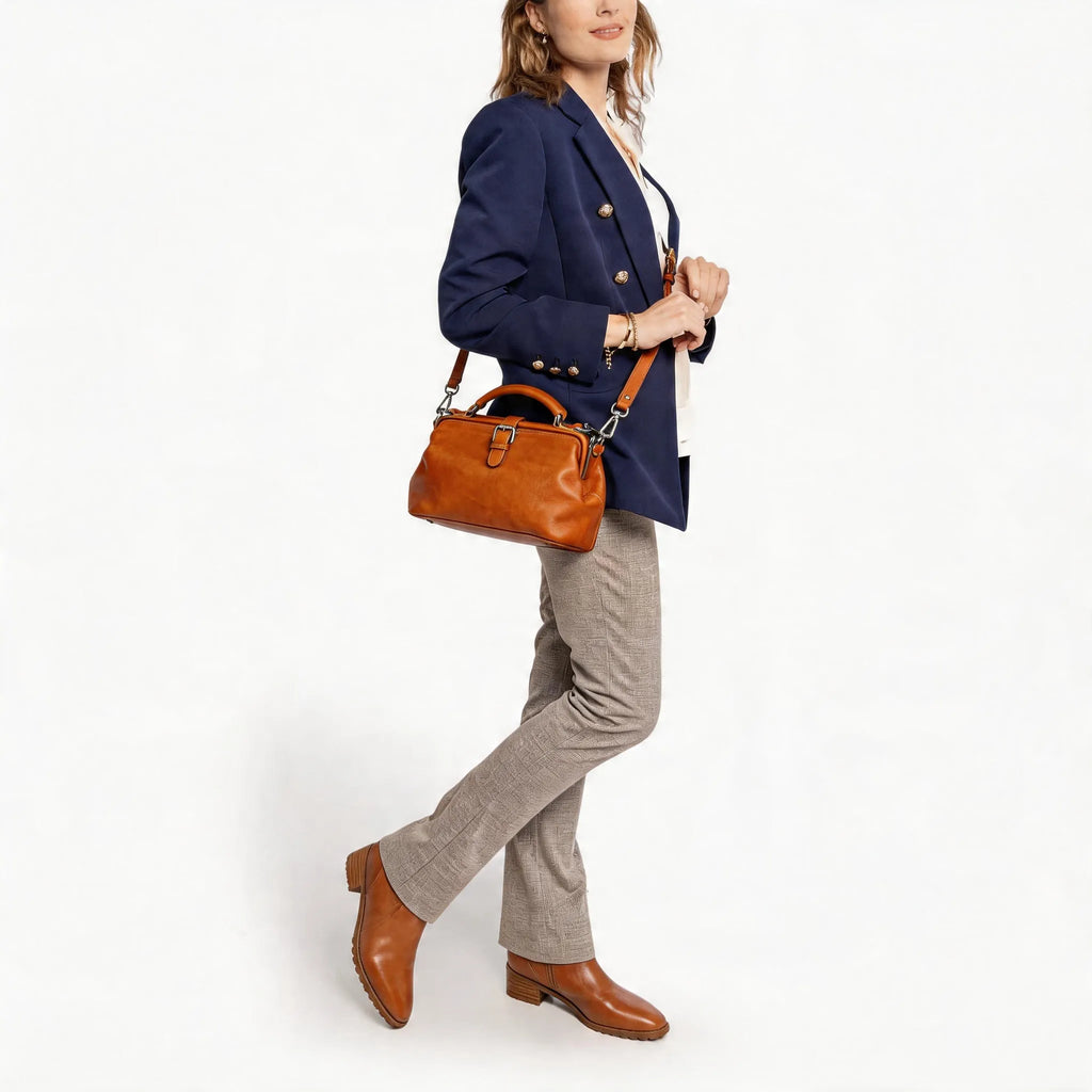 Woman in a navy blazer and gray pants holding a brown leather handbag on a white background