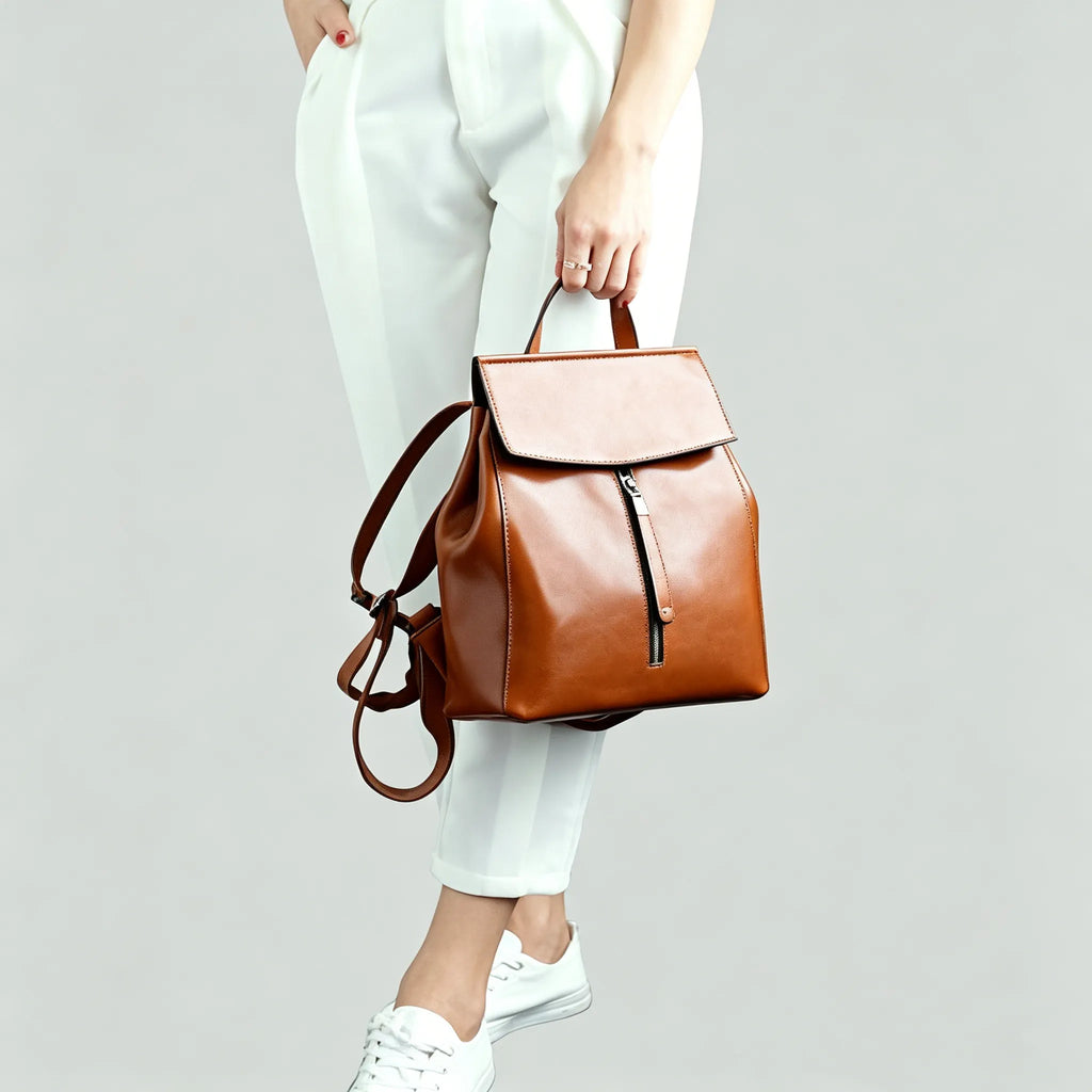 Brown leather backpack held by a person wearing white pants and shoes on a light gray background