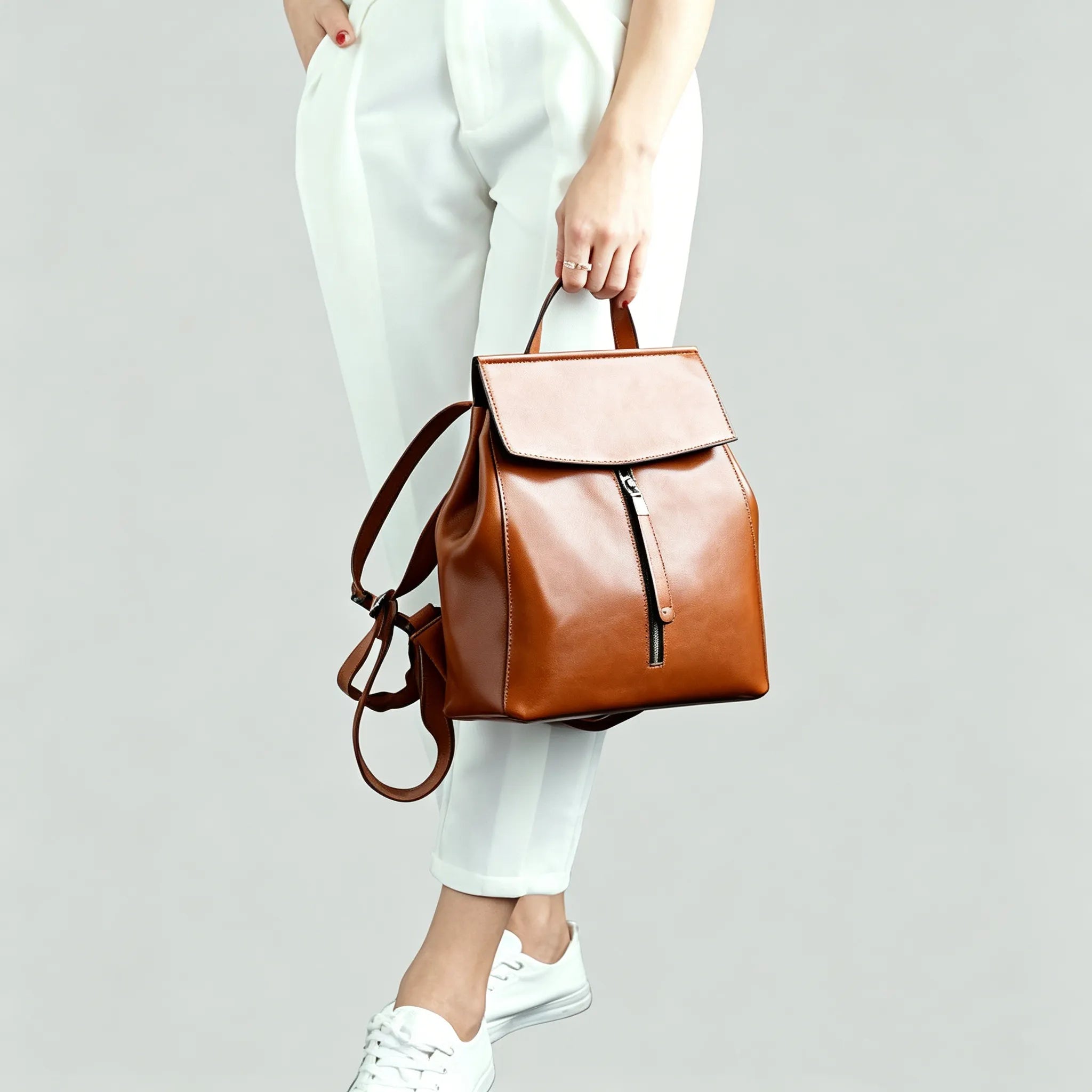 Brown leather backpack held by a person wearing white pants and shoes on a light gray background