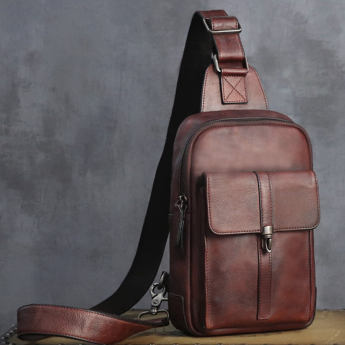 A brown leather crossbody bag with a front pocket and adjustable shoulder strap, photographed against a textured gray studio background.