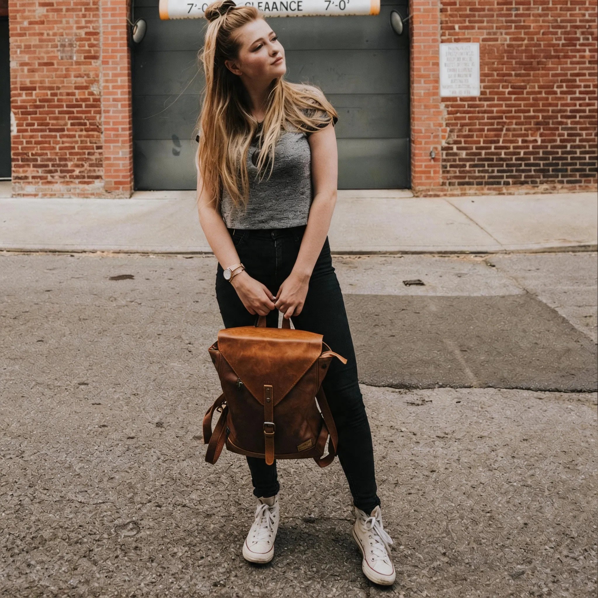 Woman holding a brown leather backpack on a street with a brick building in the background