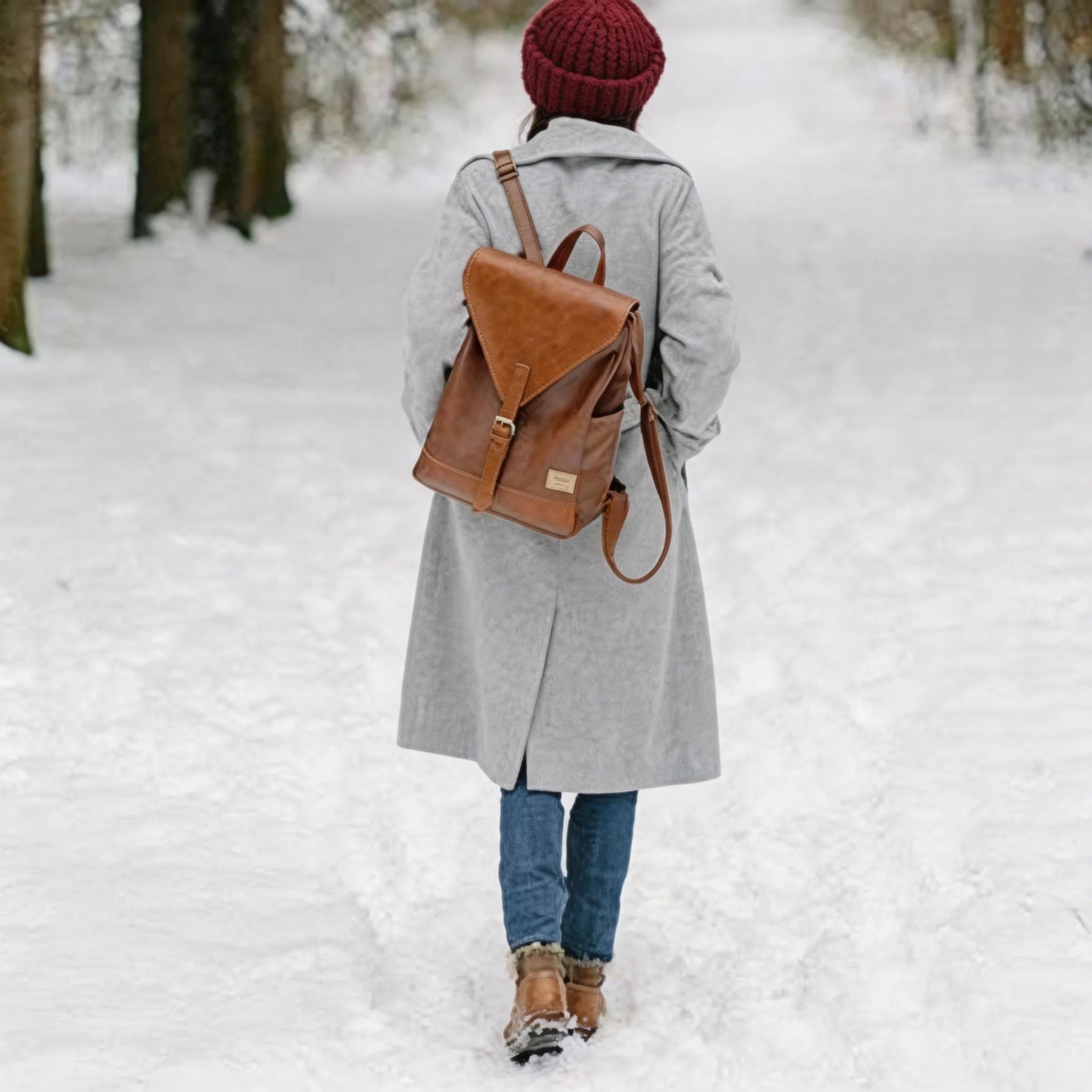 Person walking in the snow wearing a gray coat, red beanie, and brown backpack.