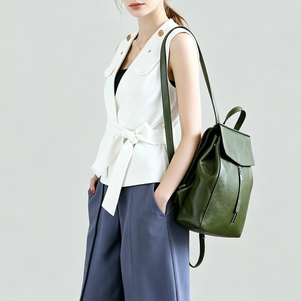 Person wearing a white sleeveless top and blue pants, holding a green leather backpack against a light gray background.