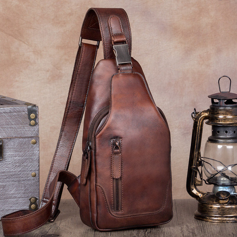 Brown leather sling bag next to a vintage suitcase and lantern on a wooden surface.