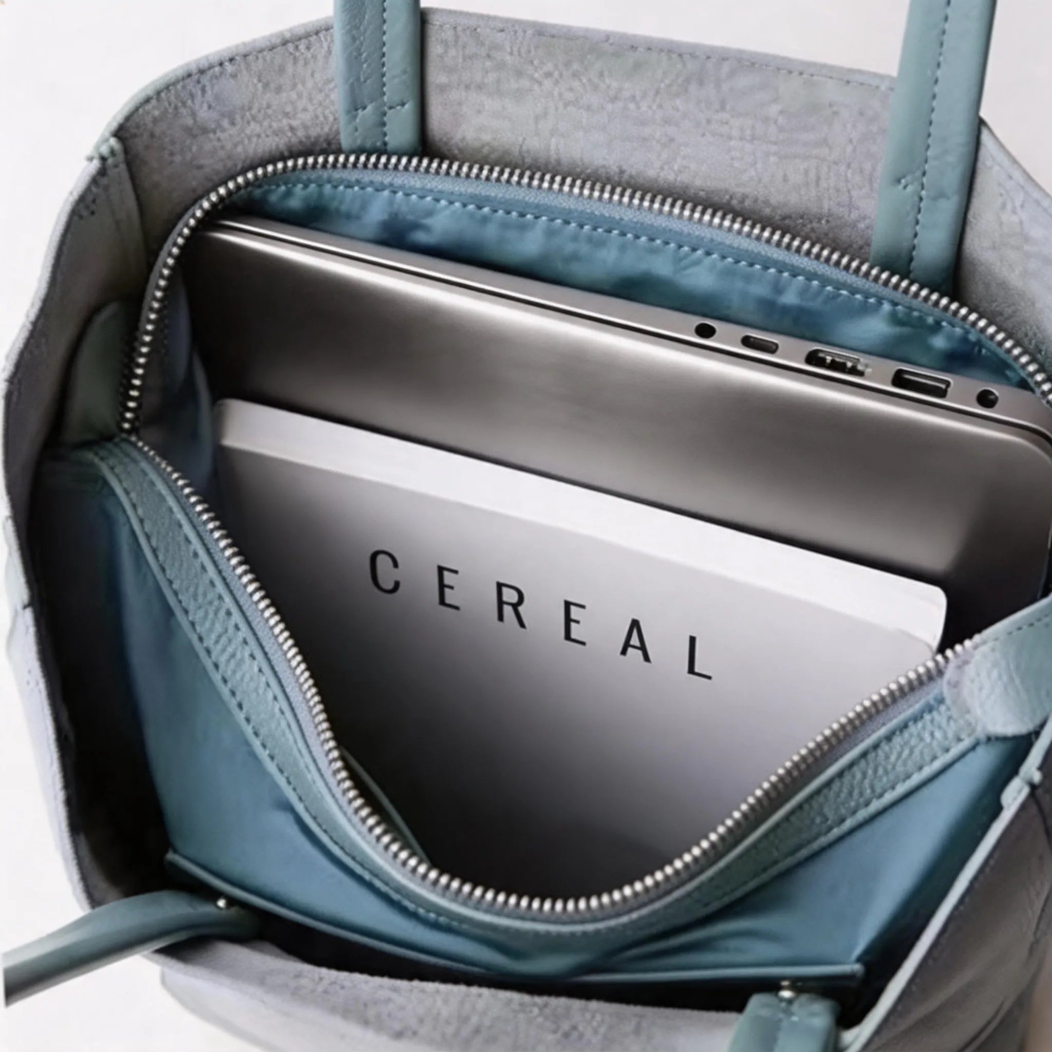 Close-up of a teal handbag with a laptop inside, featuring the brand 'CEREAL'.