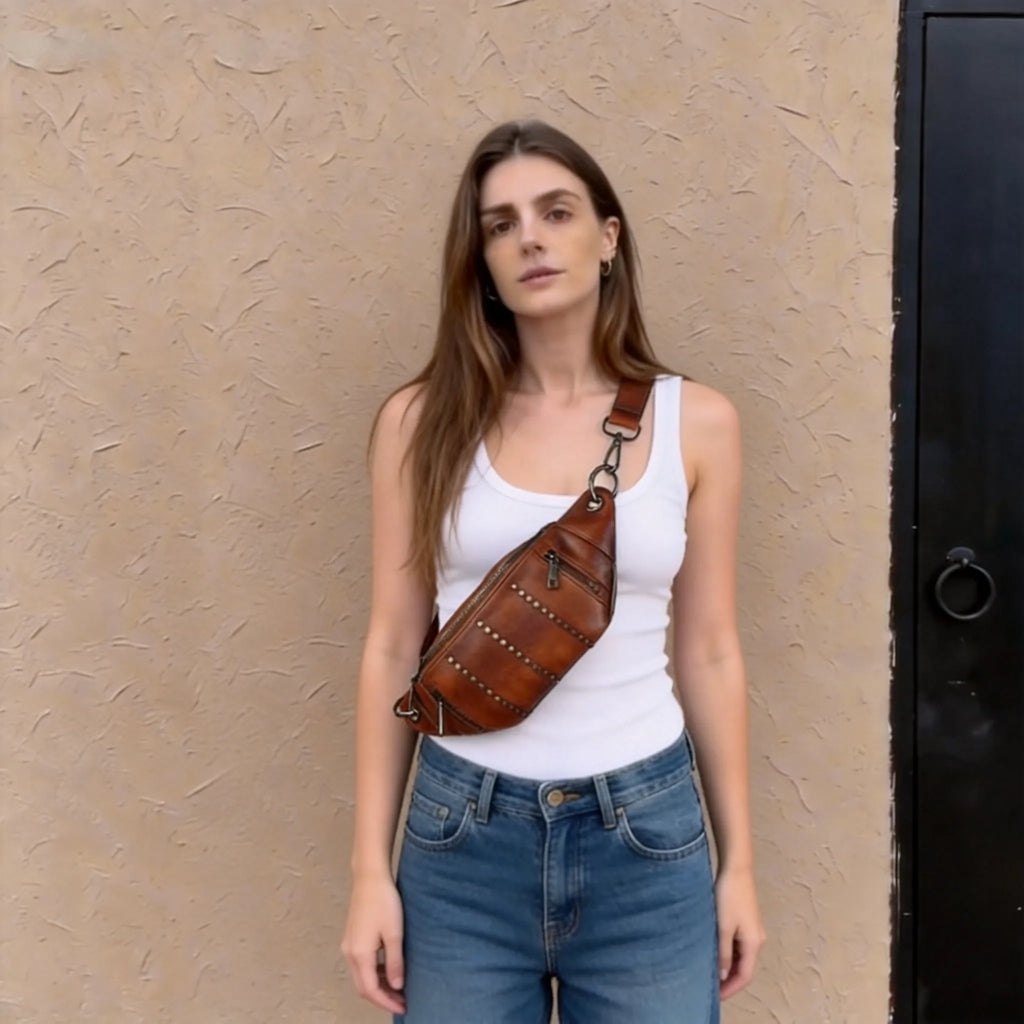 Woman wearing a white tank top and blue jeans with a brown leather bag over her shoulder against a beige wall.