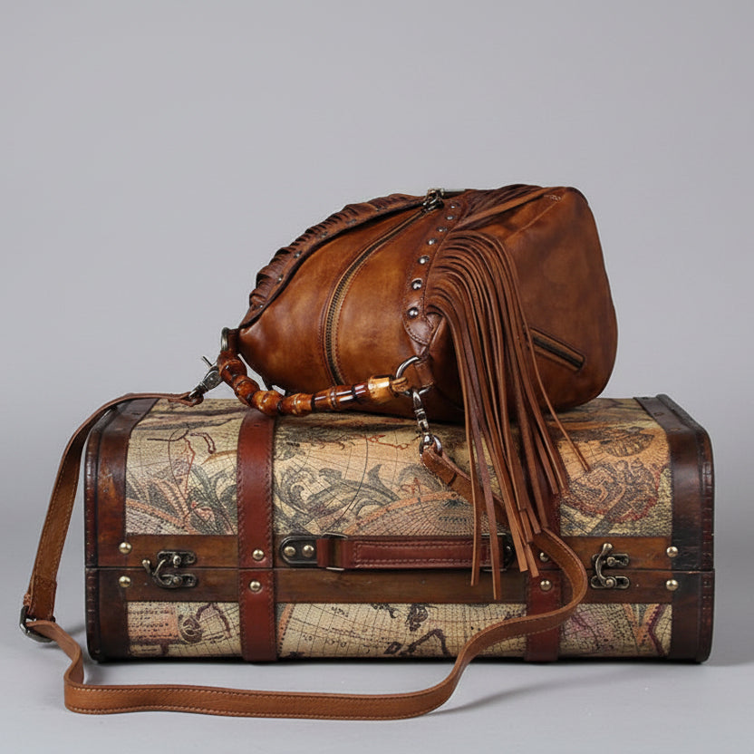 Brown leather bag with fringes on top of a vintage-style suitcase with world map design.