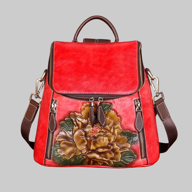 A red faux-leather backpack with brown leather straps and trim, featuring a large hand-painted peony flower motif on the front pocket. The bag is displayed on a wooden surface against a dark, textured background.