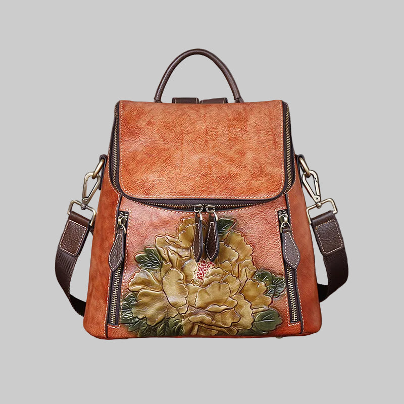 A brown faux-leather backpack with a large embossed floral design on the front pocket, featuring dark brown leather trim, top handle, and adjustable shoulder straps, shown against a solid gray background.