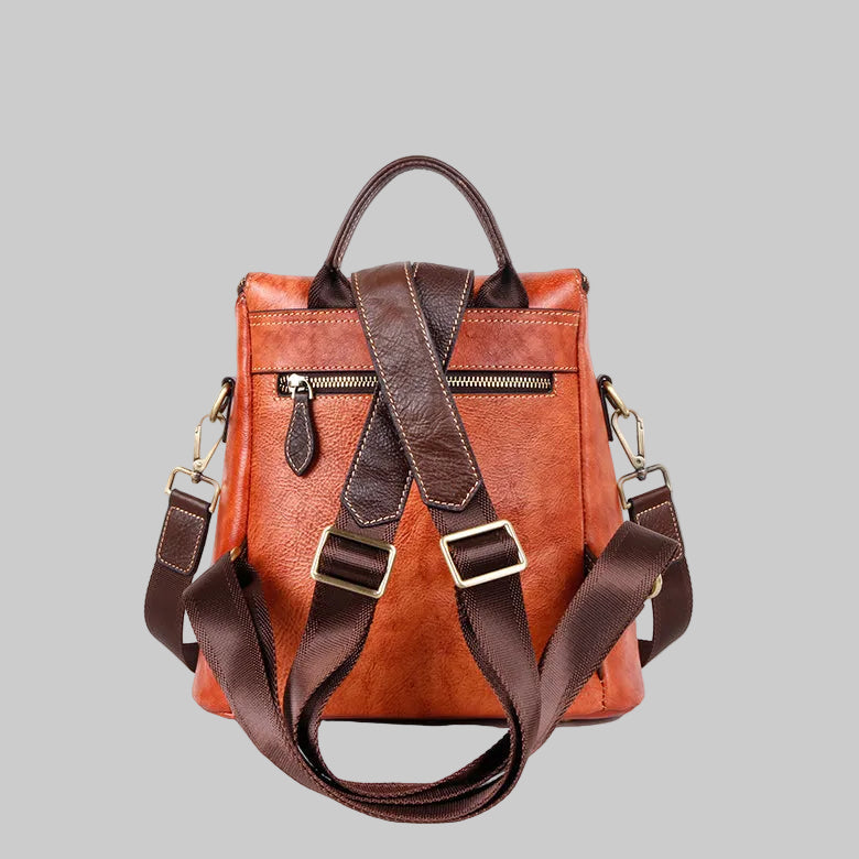 A brown leather backpack with a warm orange-brown body and dark brown straps, featuring top handles, a front zip pocket, and adjustable side straps with gold-tone metal hardware, shown against a solid gray background.