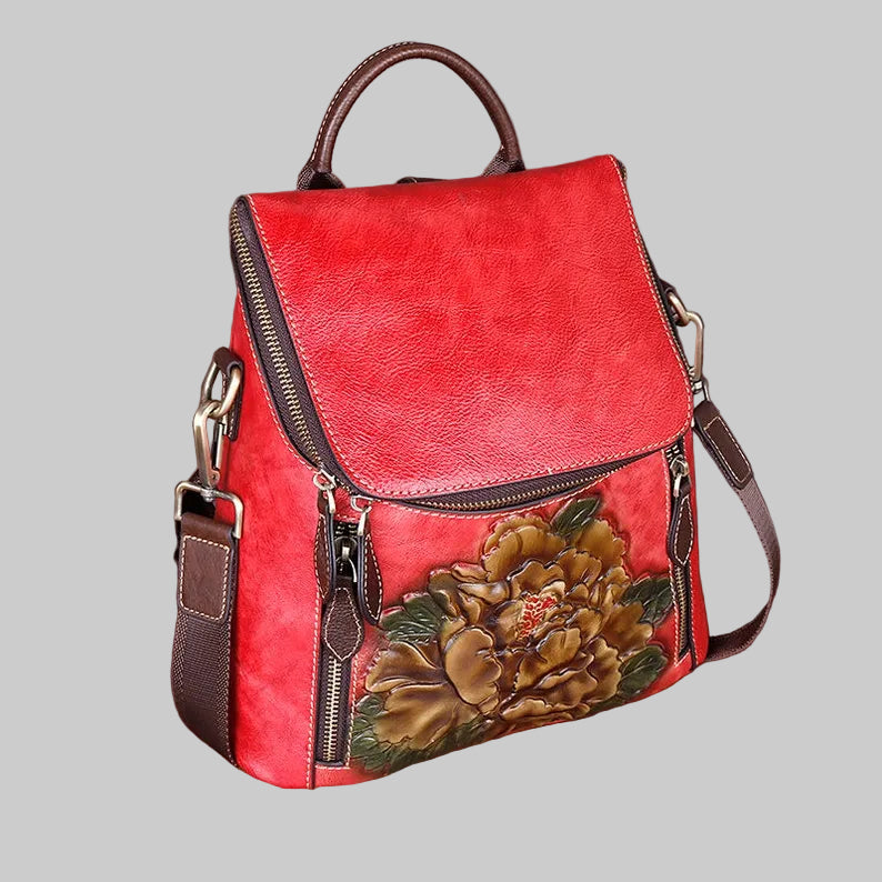 A red faux-leather backpack with a large hand-painted peony on the front, brown leather straps and handle, and gold-tone hardware, set on a wooden surface against a textured gray background.