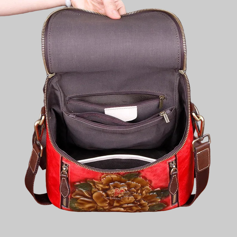 An open red shoulder bag with a large brown floral design on the front, showing a dark gray interior with multiple zippered compartments. A hand is holding the top edge of the bag against a plain light gray background.
